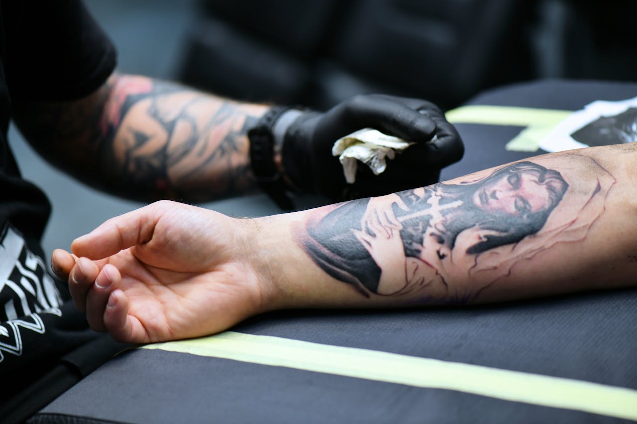 about-01 A tattoo artist creating detailed artwork on a person's arm in an indoor studio setting.