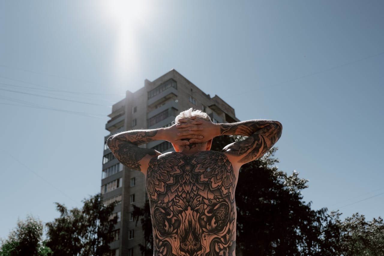 Back view of a tattooed person standing outdoors near an urban building under clear skies.
