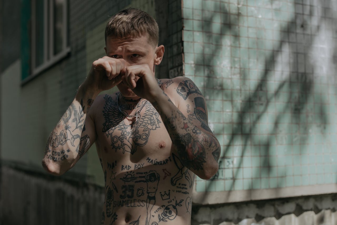 about-02 A tattooed man practices boxing techniques against a concrete wall outdoors.